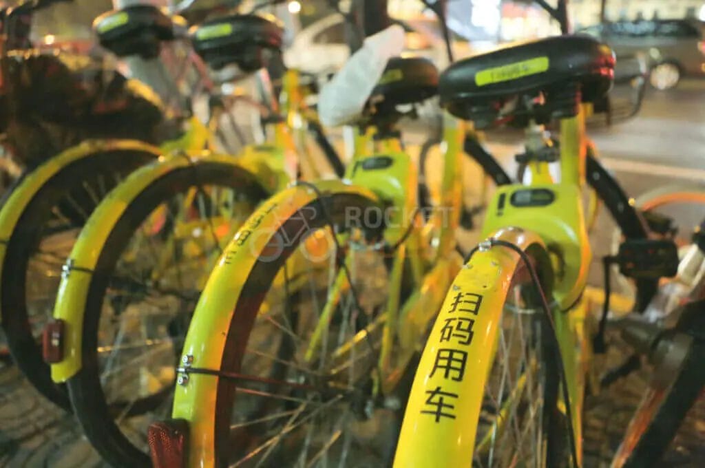 A row of yellow shared bicycles parked on a city street at night.