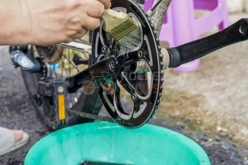 Bicycle maintenance in progress with a focus on cleaning the chain and gears using a brush and a bucket of soapy water.