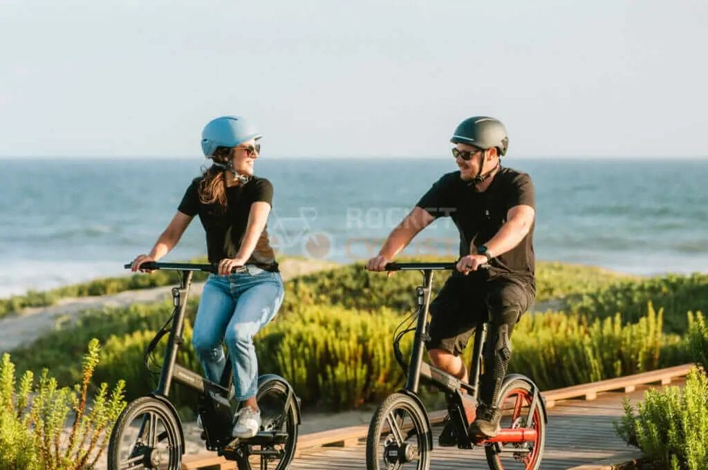 Two people riding electric bikes on a boardwalk near the ocean.