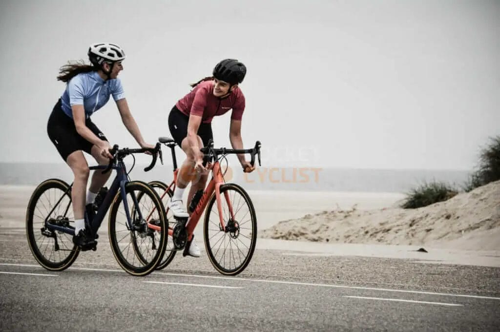 Two women riding bikes on a road near the ocean.