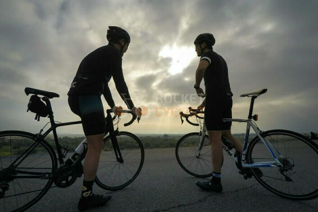 Two cyclists standing on the side of a road under a cloudy sky.