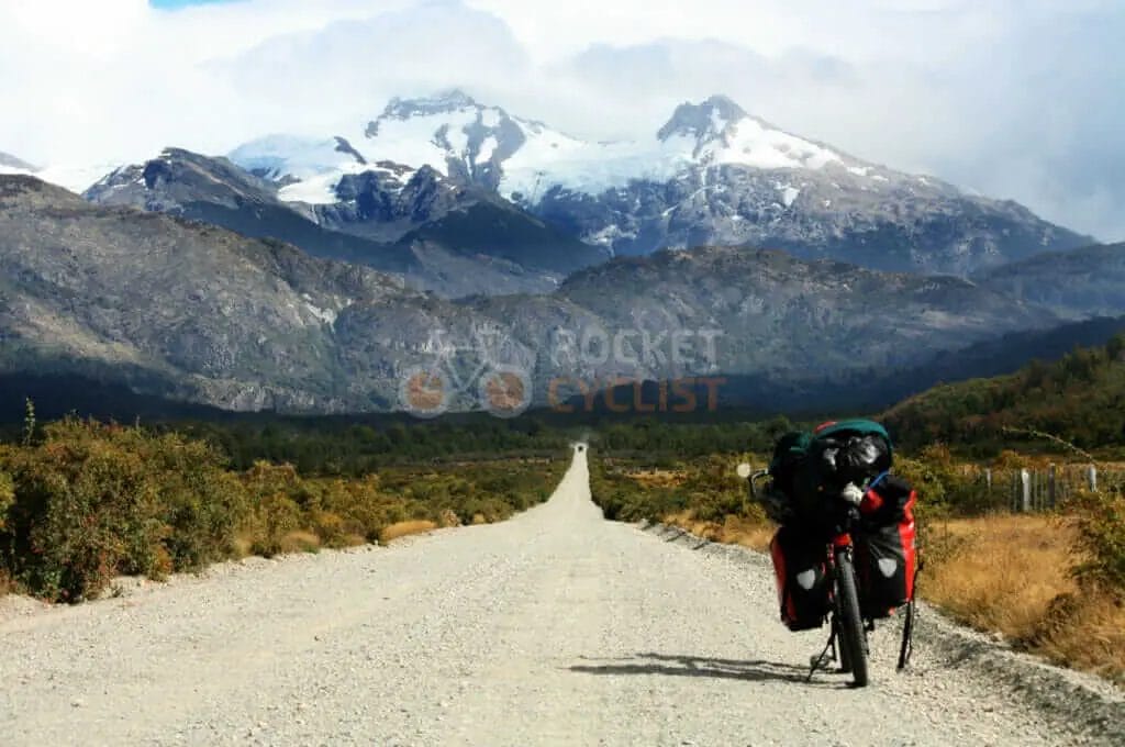 A man riding a bike down a dirt road with mountains in the background.