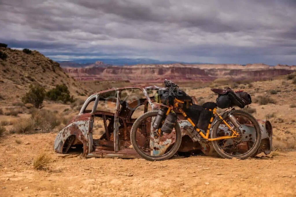 A bicycle sits next to a rusty car in the desert.