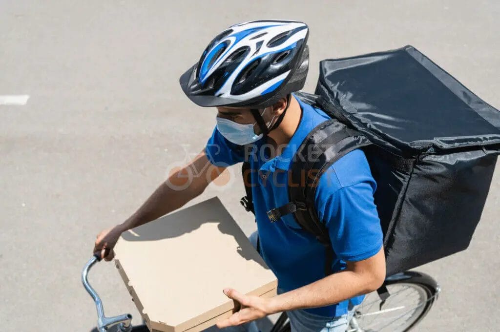 A man wearing a helmet and carrying a pizza box on a bicycle.