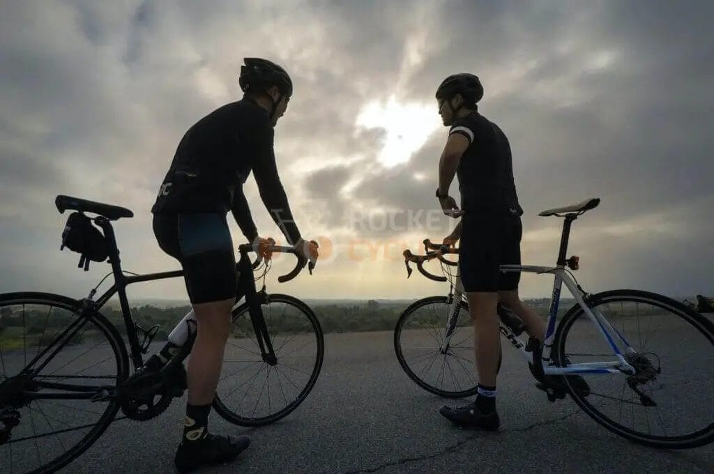 Two cyclists standing on the side of a road with a cloudy sky.