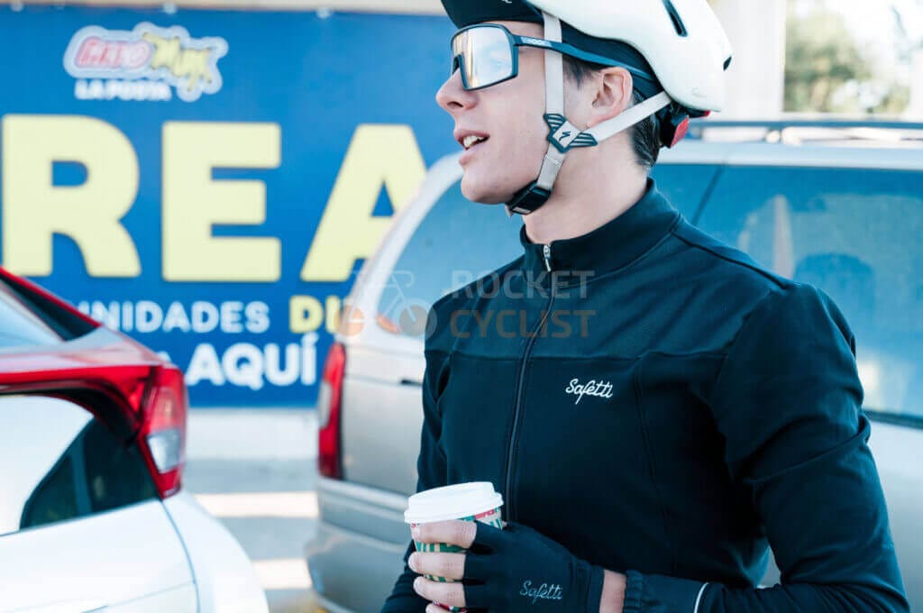 A man wearing a helmet and a cup of coffee in front of a car.