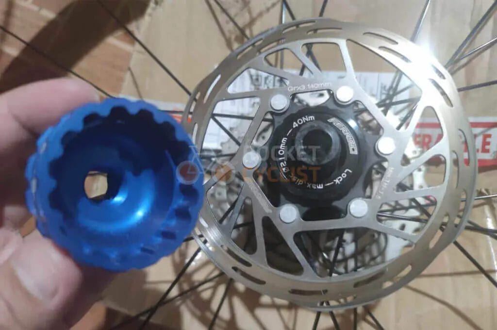 A hand holding a blue bicycle hub in focus, with a disc brake rotor on a wheel in the background.