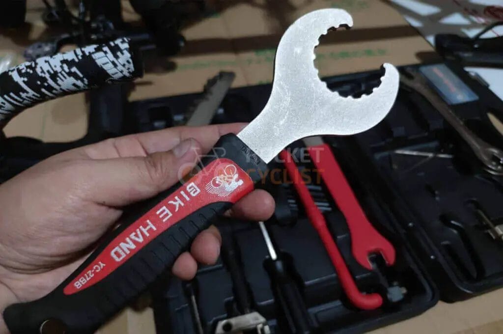 A person's hand holding a bicycle pedal wrench with a toolbox in the background.