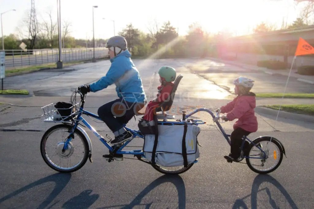 A family on a tandem bicycle with a child trailer attachment cycling in the sunlight.