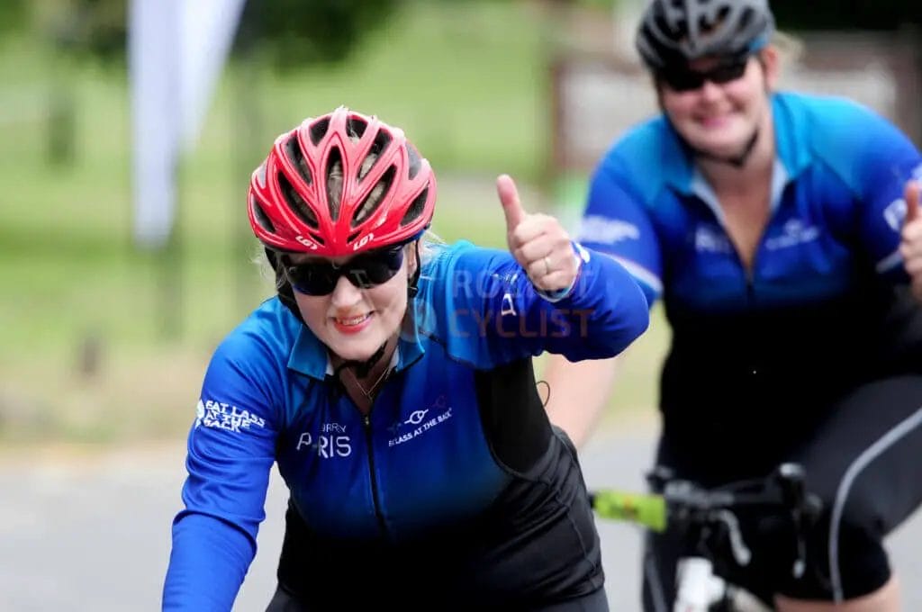 Two cyclists in blue jerseys and helmets, one gesturing with a thumbs-up.