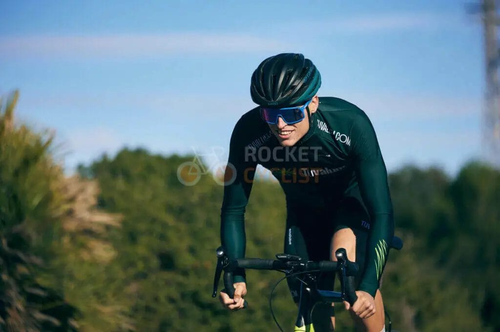A cyclist in sportswear and helmet riding on a road with vegetation in the background.