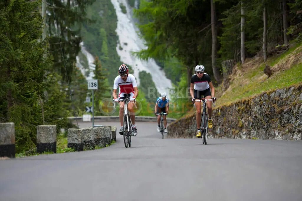 Two cyclists riding down a road near a waterfall.