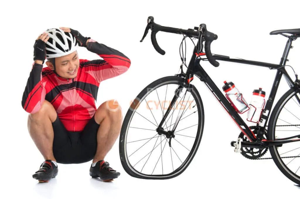 Cyclist in red and black attire sitting on the floor next to a road bike, adjusting his helmet.