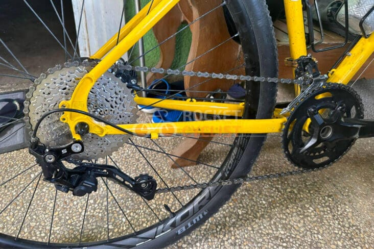 Close-up of a yellow bicycle's rear drivetrain, featuring the cassette, chain, derailleur with goatlink for smoother shifting, and crankset positioned on a textured ground surface.