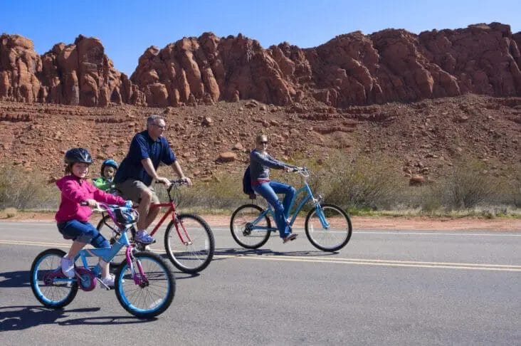 A group of four people, including two children and two adults, riding bicycles on a paved road with red rock formations in the background. Celebrating National Bicycle Day, they enjoy the ride as a perfect activity for wellness amidst stunning scenery.
