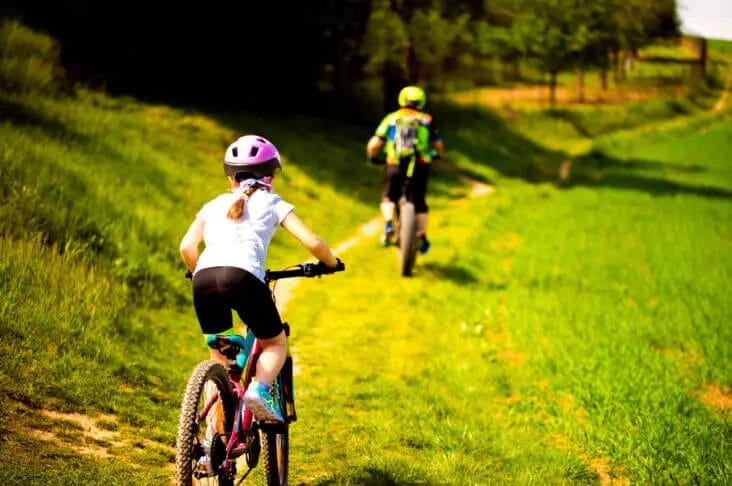 Two cyclists, one wearing a pink helmet, ride along a grassy trail surrounded by trees and greenery on a sunny day, showcasing the simplicity of their Microshift Acolyte groupset.