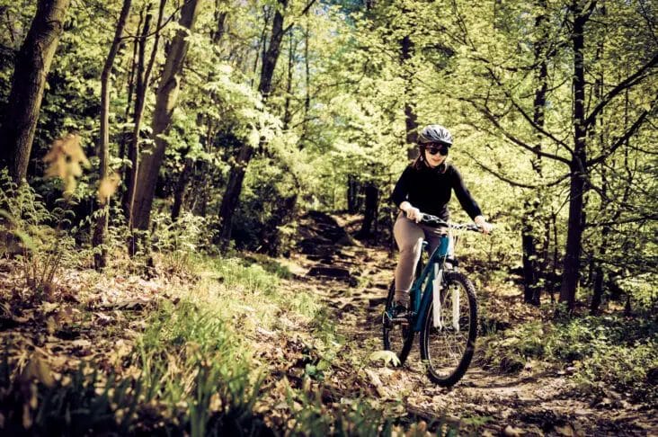 A person wearing a helmet navigates off-road on a mountain bike along a forest trail surrounded by lush green trees.