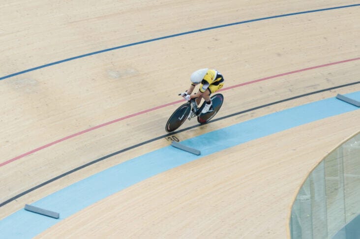 Experience a cyclist in yellow and white gear riding on an indoor velodrome track, angled towards a corner; it's truly a cyclist's dream.